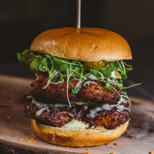 Juicy double chicken burger with BBQ rub, melted cheese, microgreens, and pickles on a wooden board