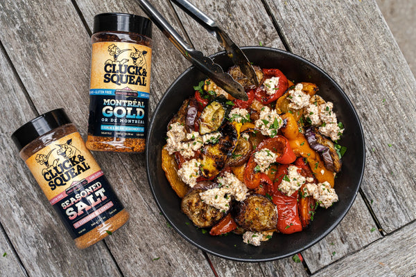Grilled vegetables with feta in black bowl beside Cluck & Squeal Montreal Gold and Seasoned Salt rubs on wood table